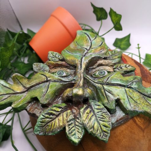 Clay greenman laid on a wooden block surrounded by ivy and a turned over terracotta plant pot.