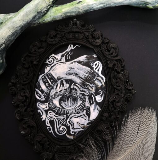 A photo of a black and white drawing of a mystical looking eye, with a monster hand over the brow. Drawing is in a black oval baroque esq style frame.